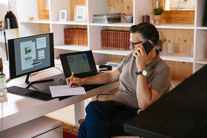 Middle aged man in casual clothes and glasses talking on cellphone and writing in planner while sitting at desk and working on design project in home office.