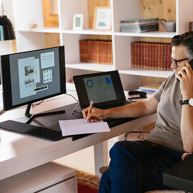 Middle aged man in casual clothes and glasses talking on cellphone and writing in planner while sitting at desk and working on design project in home office.